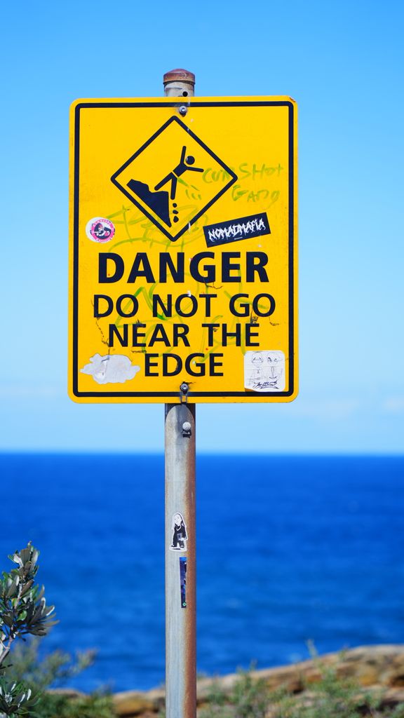 Yellow warning sign with graffiti by ocean, advising caution near the edge.